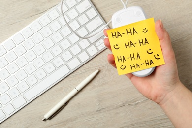 Woman holding computer mouse with sticky note at wooden table, top view. April fool's day