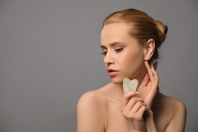 Beautiful young woman doing facial massage with gua sha tool on grey background. Space for text