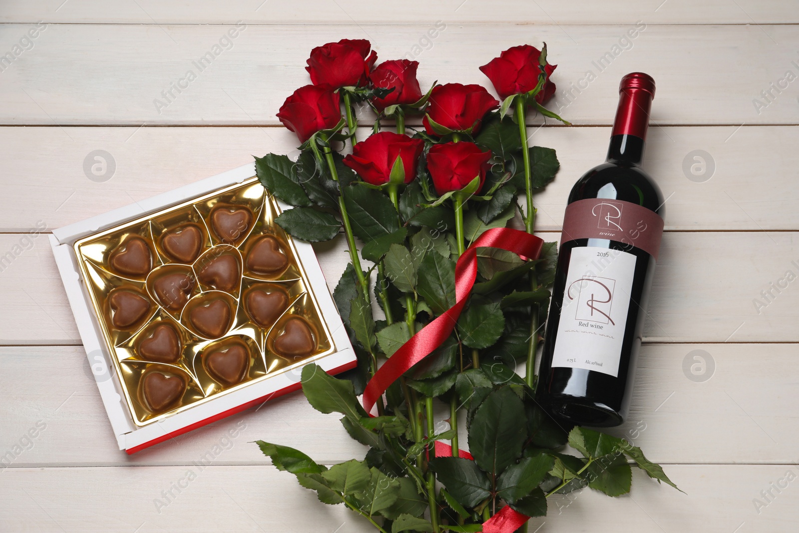 Photo of Bottle of red wine, beautiful roses and heart shaped chocolate candies on white wooden table, flat lay