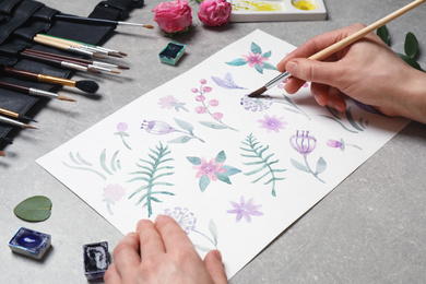 Photo of Woman painting flowers with watercolor at grey stone table, closeup