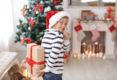 Photo of Cute little child in Santa hat hiding Christmas gift box behind back at home