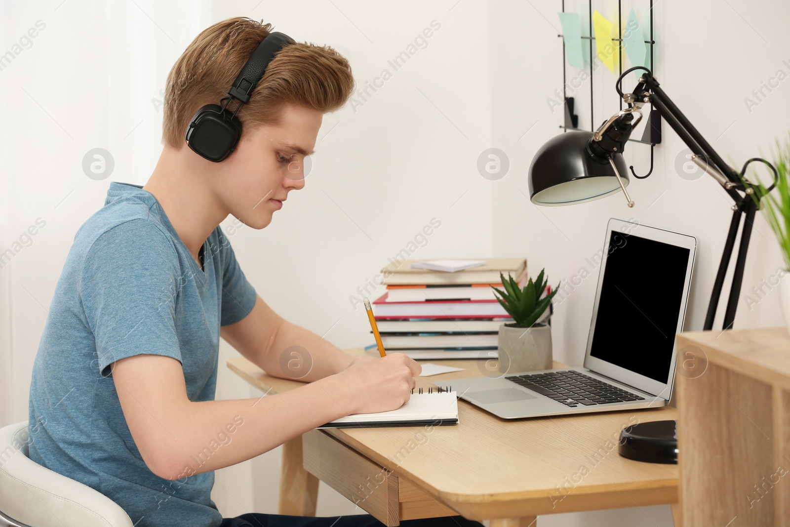 Photo of Teenage boy with headphones writing in notebook at wooden table indoors