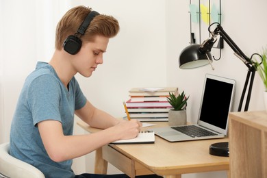 Photo of Teenage boy with headphones writing in notebook at wooden table indoors