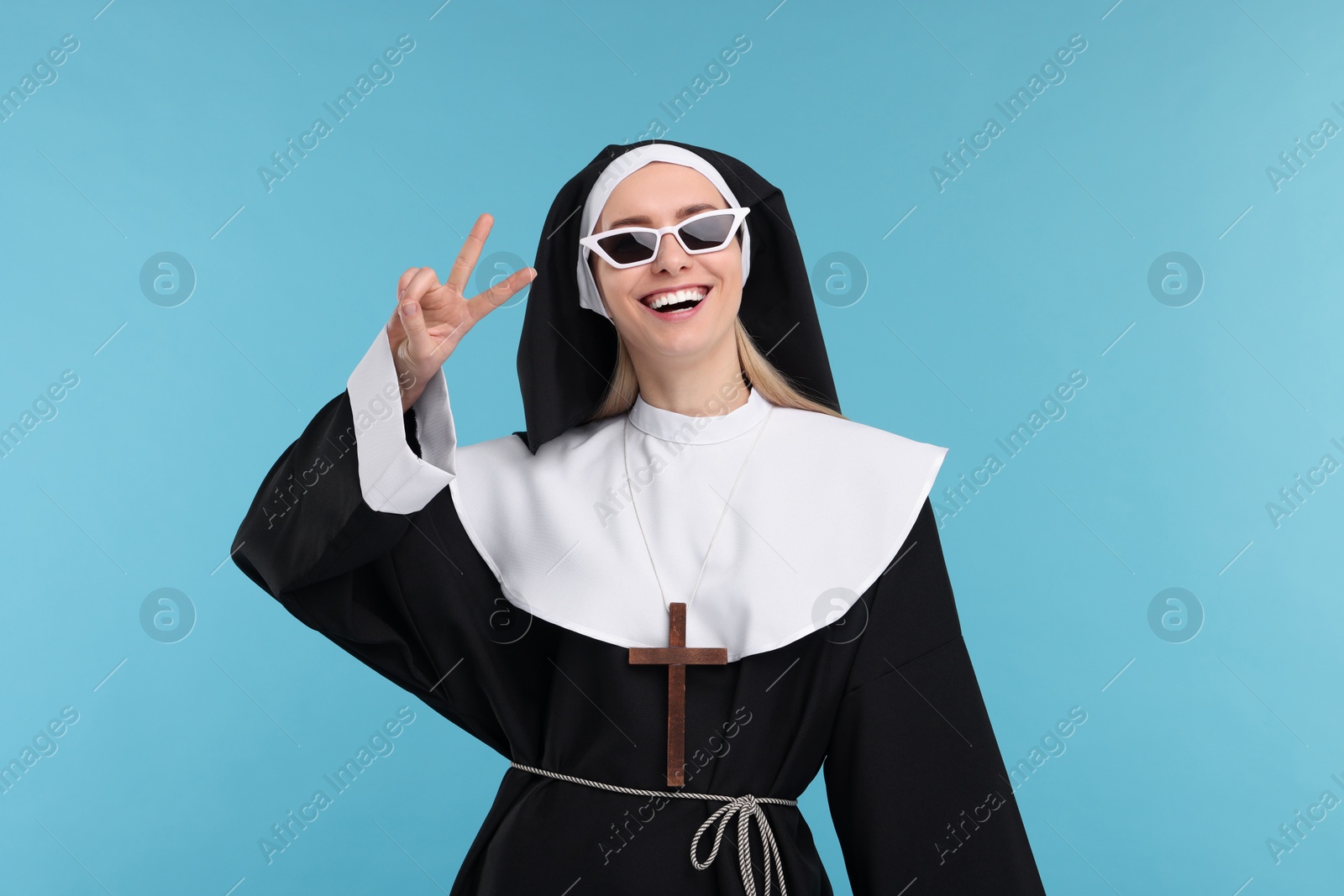 Photo of Happy woman in nun habit showing V-sign against light blue background
