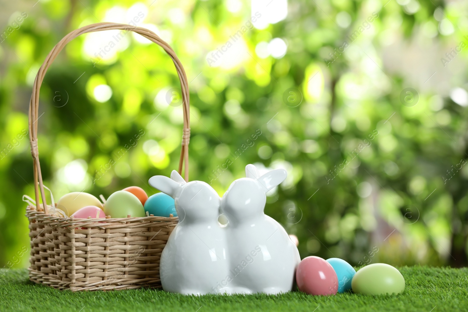 Photo of Cute ceramic Easter bunnies with wicker basket and dyed eggs on green grass against blurred background, space for text