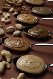 Photo of Tasty nut butters in spoons and raw nuts on brown table, closeup
