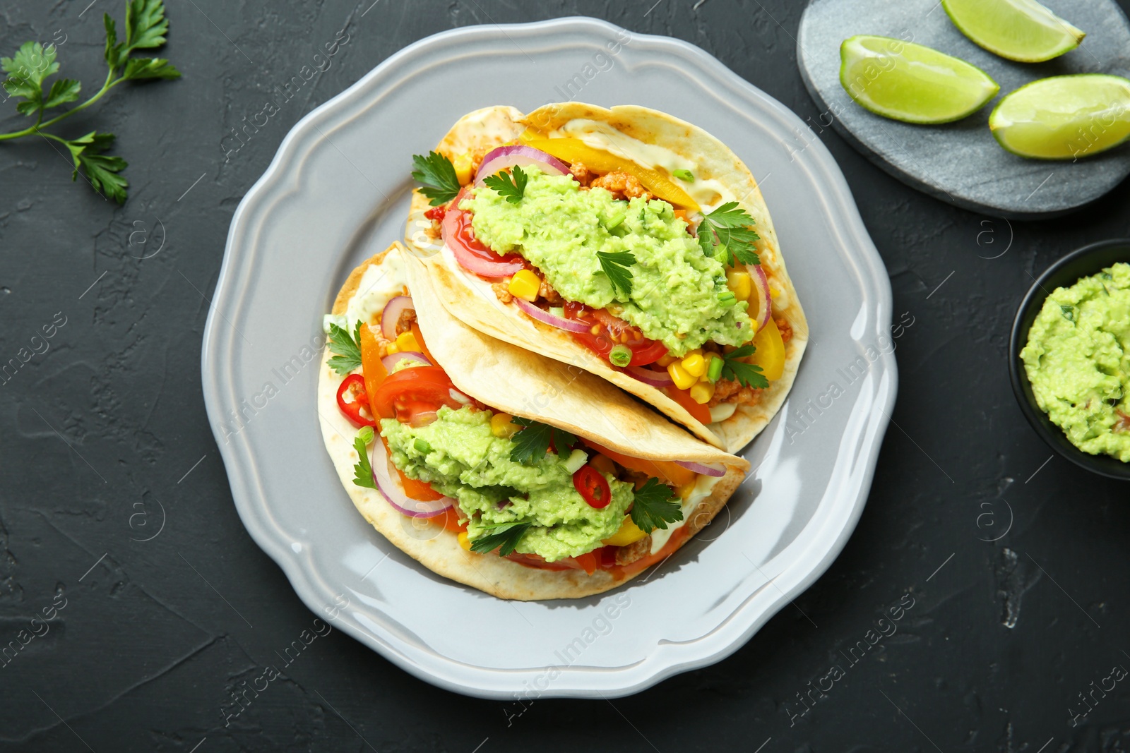 Photo of Delicious tacos with guacamole and vegetables served on black table, flat lay