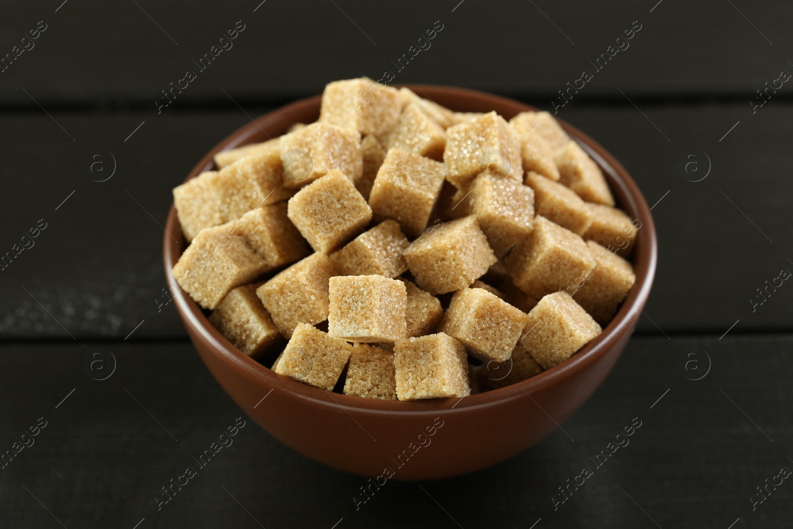 Photo of Brown sugar cubes in bowl on black wooden table, closeup