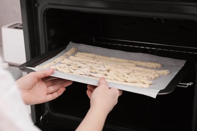 Woman putting baking sheet with homemade breadsticks into oven in kitchen, closeup. Cooking traditional grissini