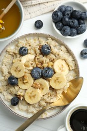 Tasty oatmeal with banana, blueberries, walnuts and honey served in bowl on white wooden table, flat lay