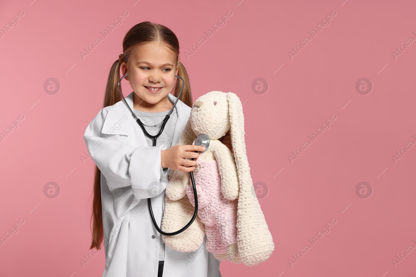 Photo of Little girl in medical uniform examining toy bunny with stethoscope on pink background. Space for text