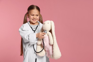 Little girl in medical uniform examining toy bunny with stethoscope on pink background. Space for text