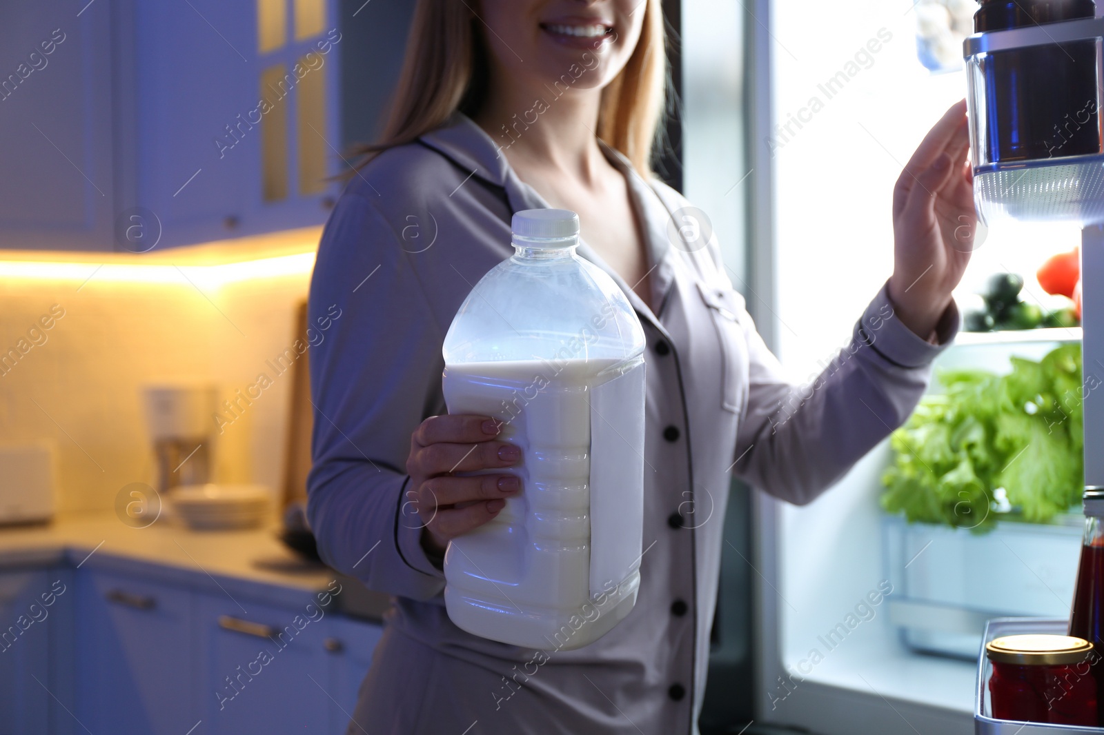 Photo of Young woman holding gallon bottle of milk near refrigerator in kitchen at night, closeup