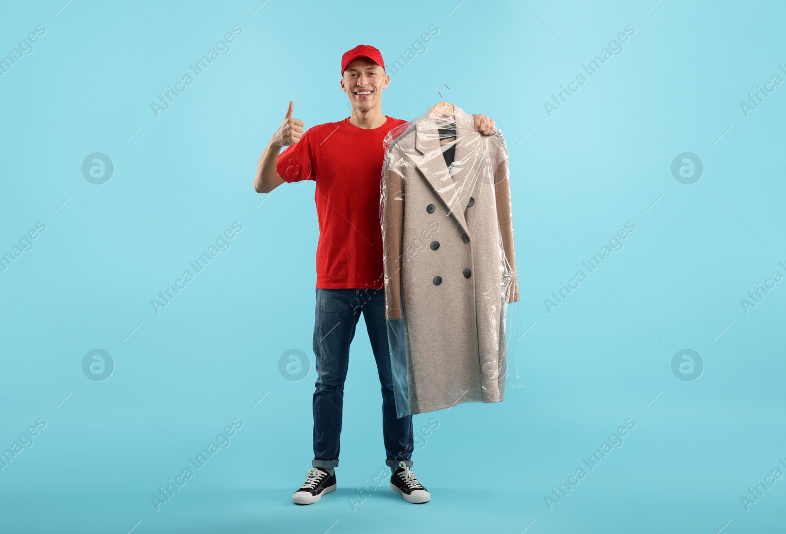 Photo of Dry-cleaning delivery. Happy courier holding coat in plastic bag and showing thumbs up on light blue background