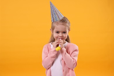 Birthday celebration. Cute little girl in party hat with blower on orange background
