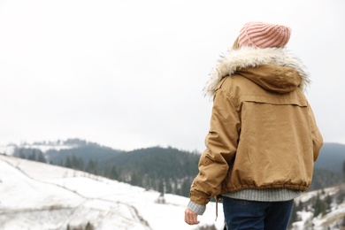 Young woman in warm clothes near snowy hill, space for text. Winter vacation