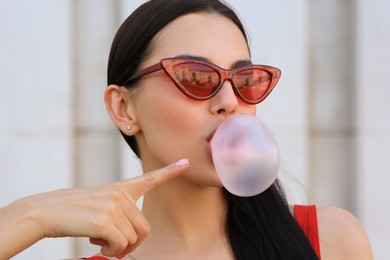 Beautiful woman in stylish sunglasses blowing gum near wall outdoors