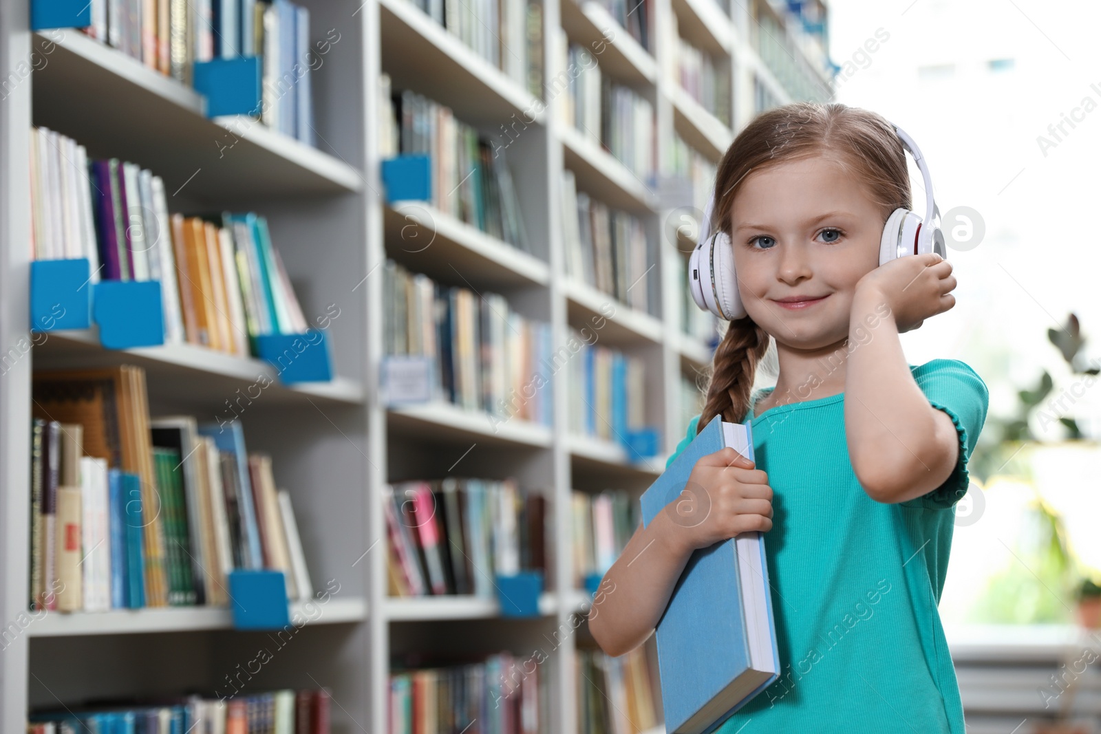 Photo of Cute little girl with headphones and book in library reading room