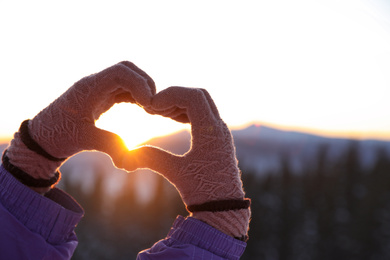 Photo of Woman making heart with her hands in mountains at sunset, closeup. Winter vacation