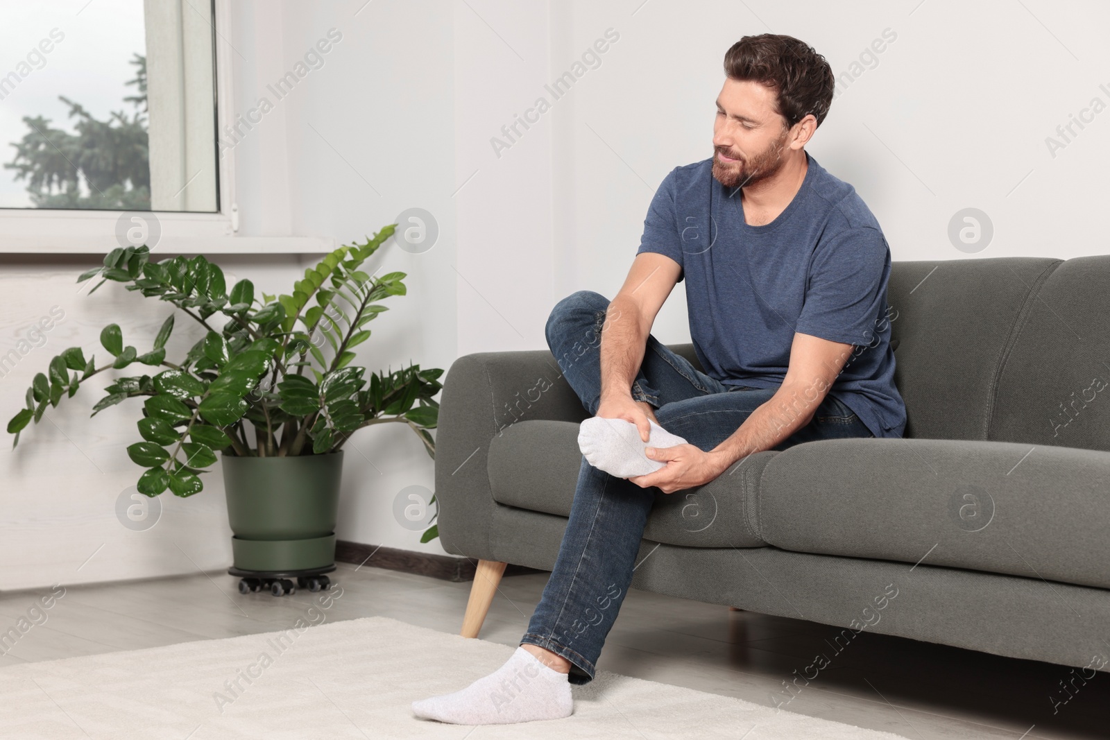 Photo of Man suffering from leg pain and massaging heel on sofa at home