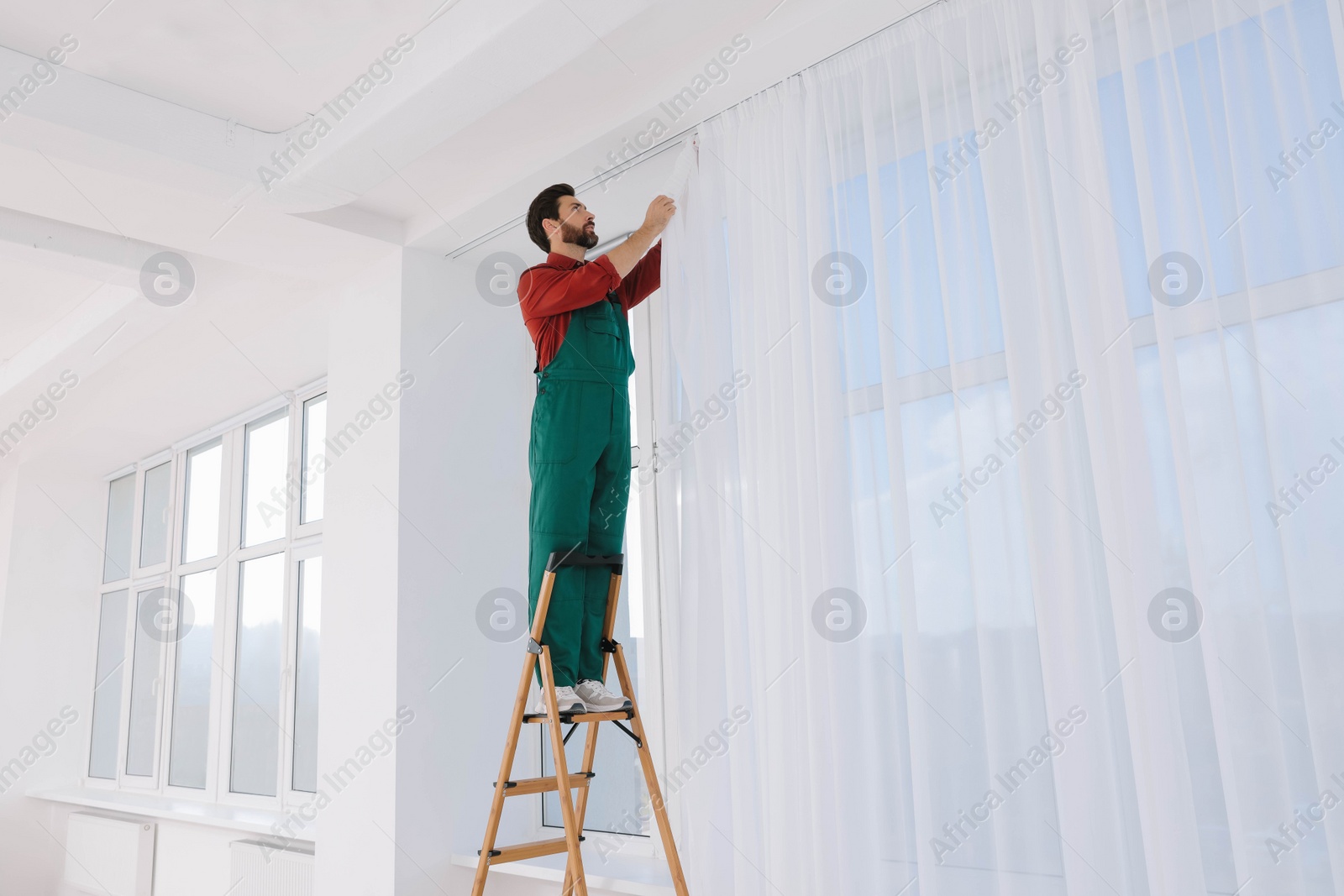Photo of Worker in uniform hanging window curtain indoors, low angle view