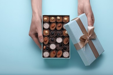 Photo of Woman with open box of delicious chocolate candies on light blue background, top view