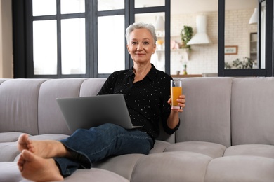 Mature woman with laptop and drink sitting on sofa at home. Smart aging