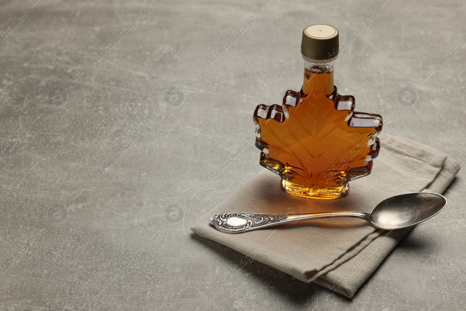 Photo of Leaf shaped bottle of tasty maple syrup and spoon on light grey table, space for text