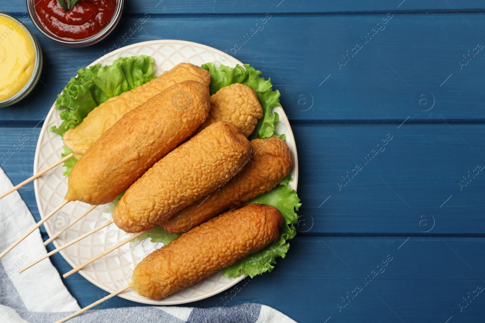 Photo of Delicious deep fried corn dogs and sauces on blue wooden table, flat lay. Space for text