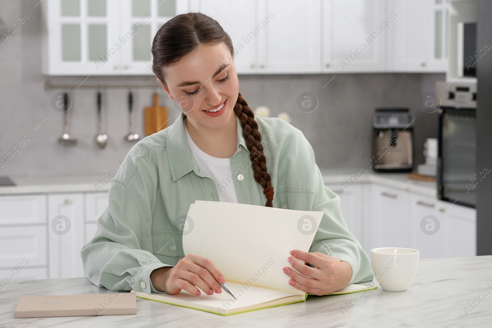 Photo of Beautiful young woman writing in notebook at white marble table indoors