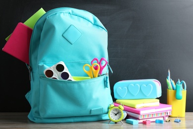 Photo of Backpack full of different school stationery on table near chalkboard