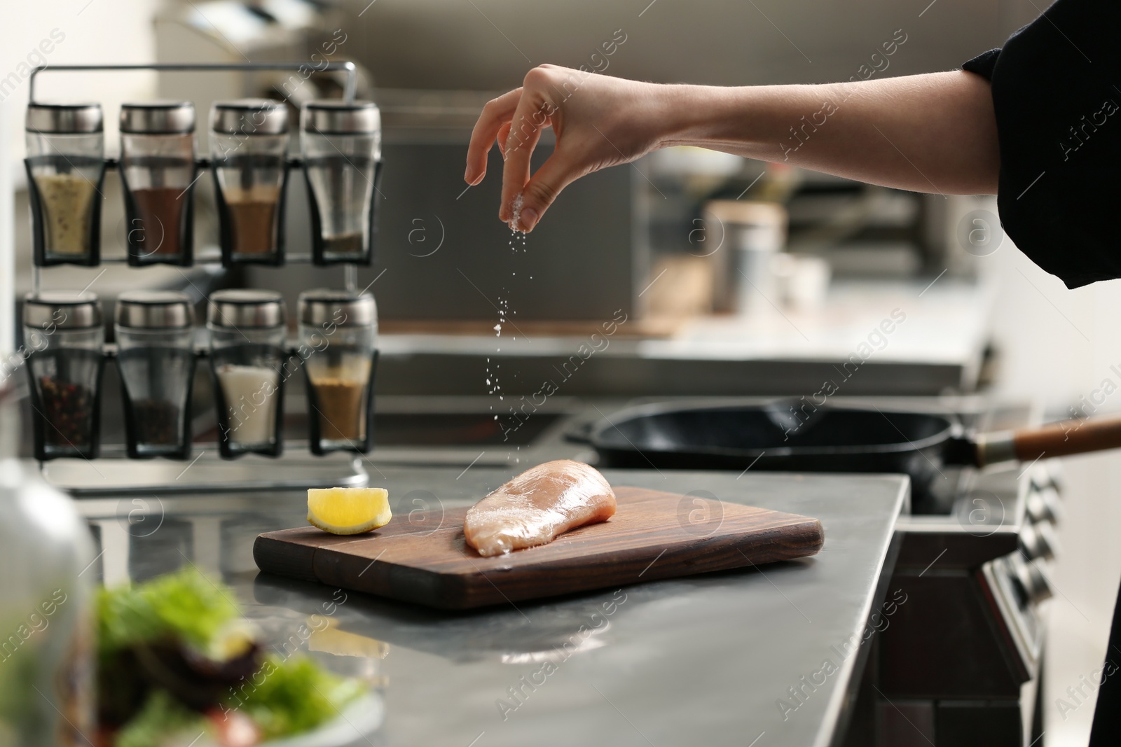 Photo of Female chef adding salt to raw chicken fillet in restaurant kitchen, closeup. Cooking food