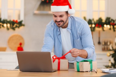 Celebrating Christmas online with exchanged by mail presents. Smiling man in Santa hat opening gift box during video call on laptop at home