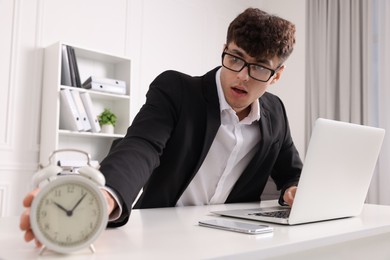 Photo of Emotional young man turning off alarm clock at white table in office. Deadline concept