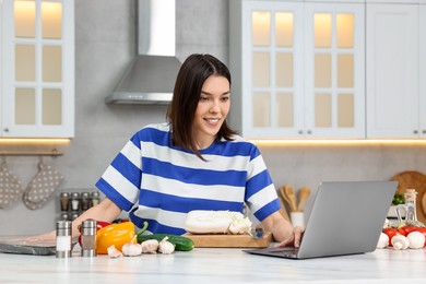 Photo of Young woman cooking while watching online course via laptop in kitchen