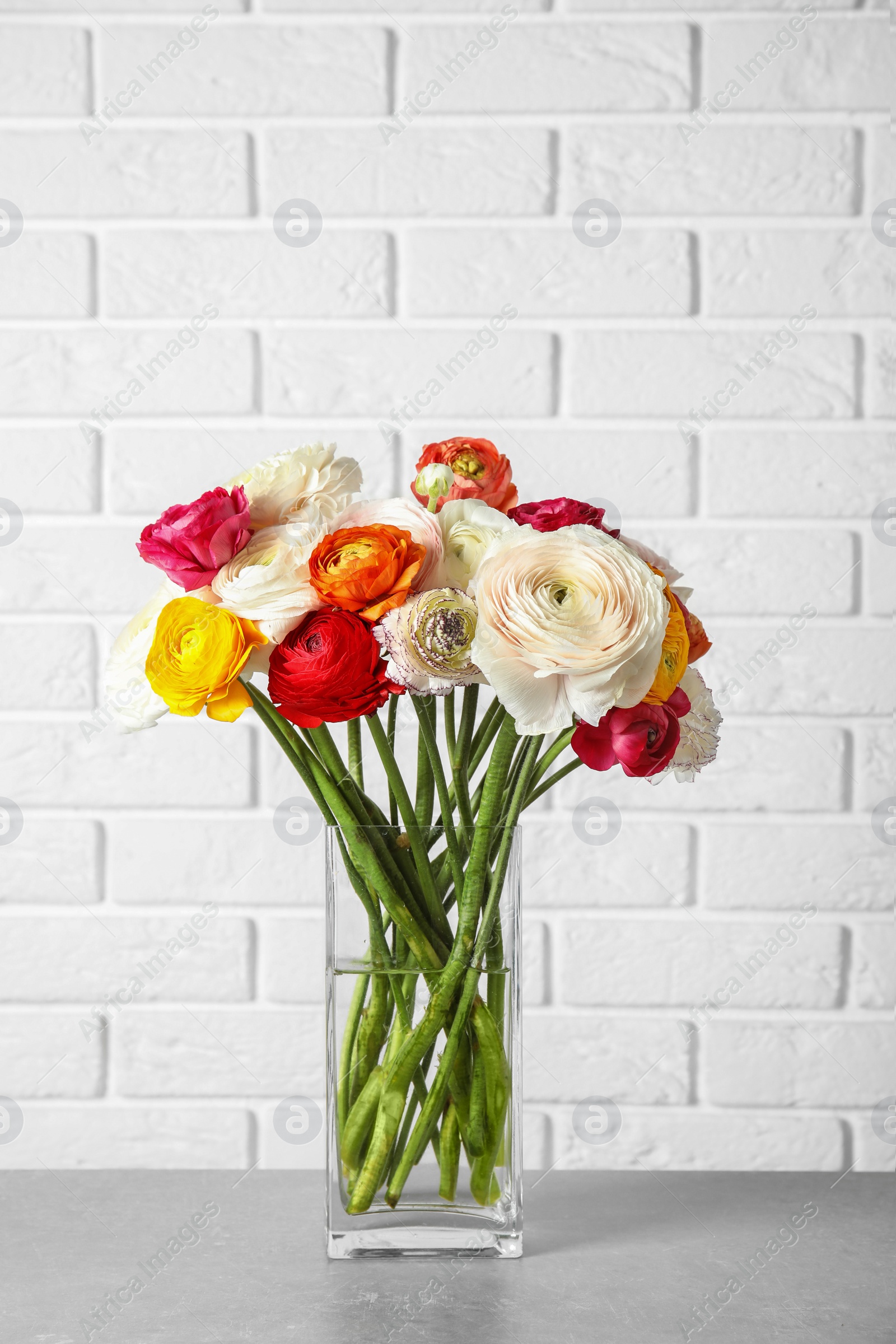 Photo of Vase with beautiful spring ranunculus flowers on table near brick wall