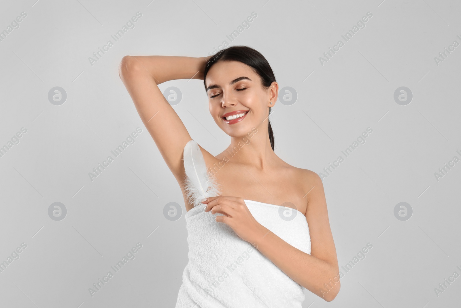 Photo of Young woman touching armpit with feather after epilation procedure on light grey background