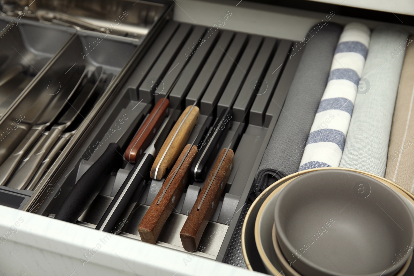Photo of Open drawer of kitchen cabinet with different utensils, dishware and towels, closeup