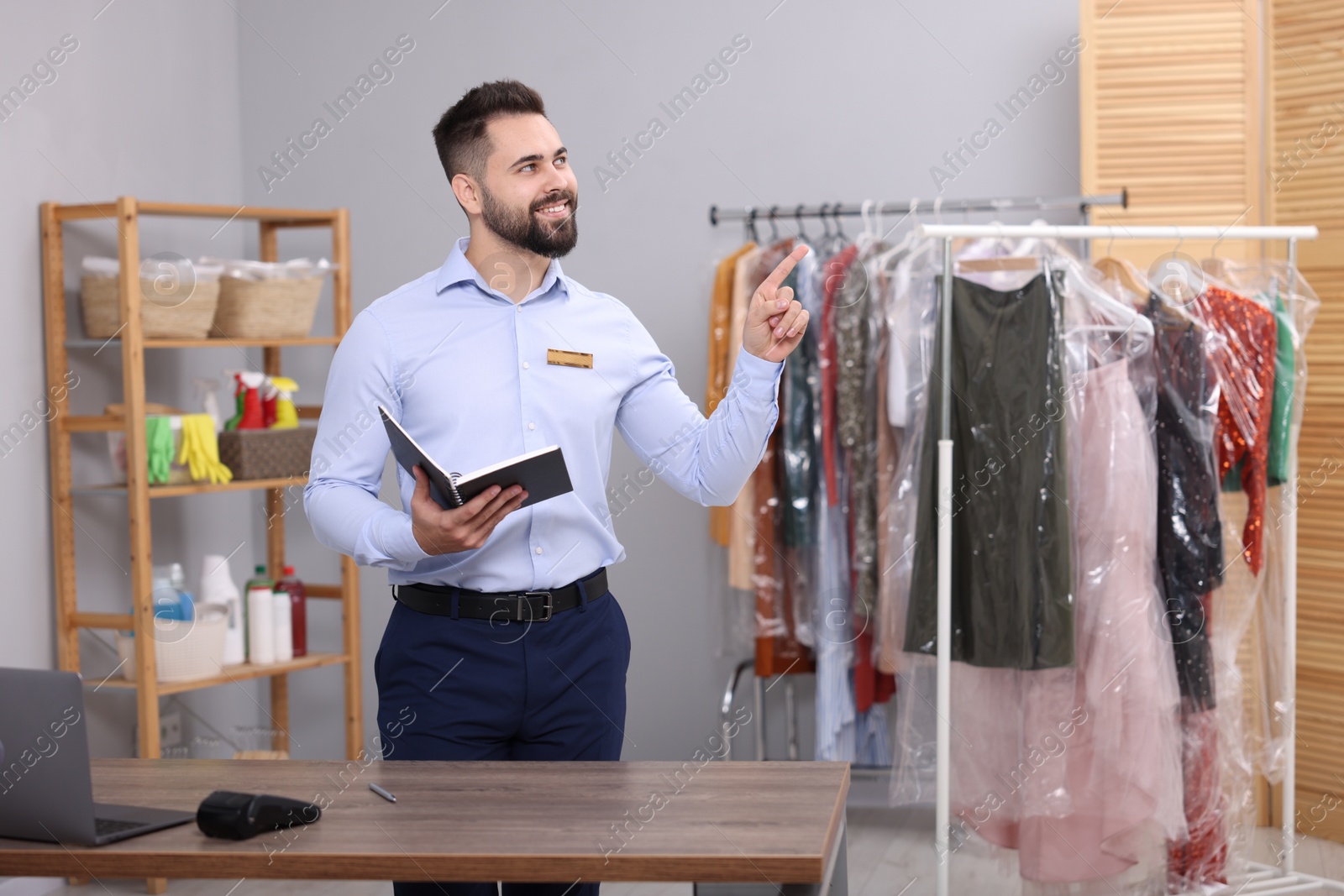 Photo of Dry-cleaning service. Happy worker with notebook pointing at something in workplace indoors, space for text