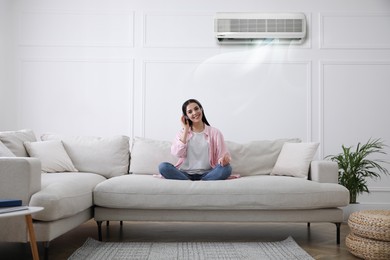 Young woman resting under air conditioner on white wall at home