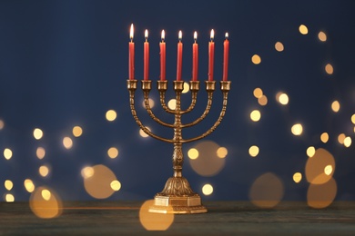 Photo of Golden menorah with burning candles on table against blue background and blurred festive lights