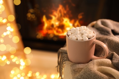 Cup of hot drink with marshmallows near fireplace at home. Space for text