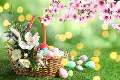 Wicker basket with Easter eggs and beautiful flowers on green grass outdoors