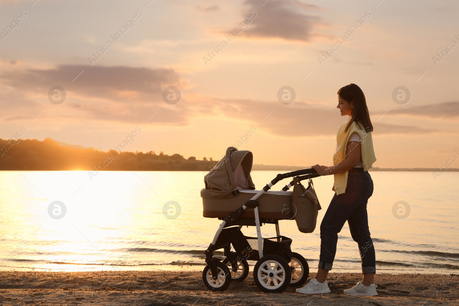 Photo of Happy mother with baby in stroller walking near river at sunset