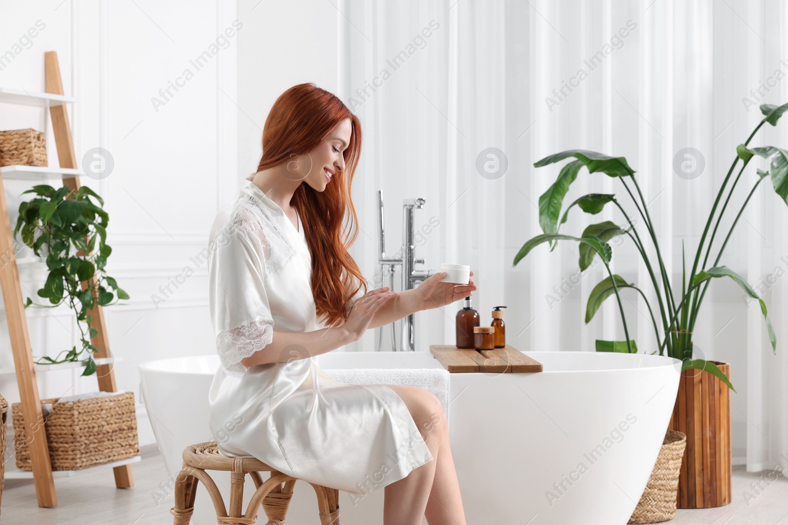 Photo of Beautiful young woman applying body cream onto arms in bathroom, space for text