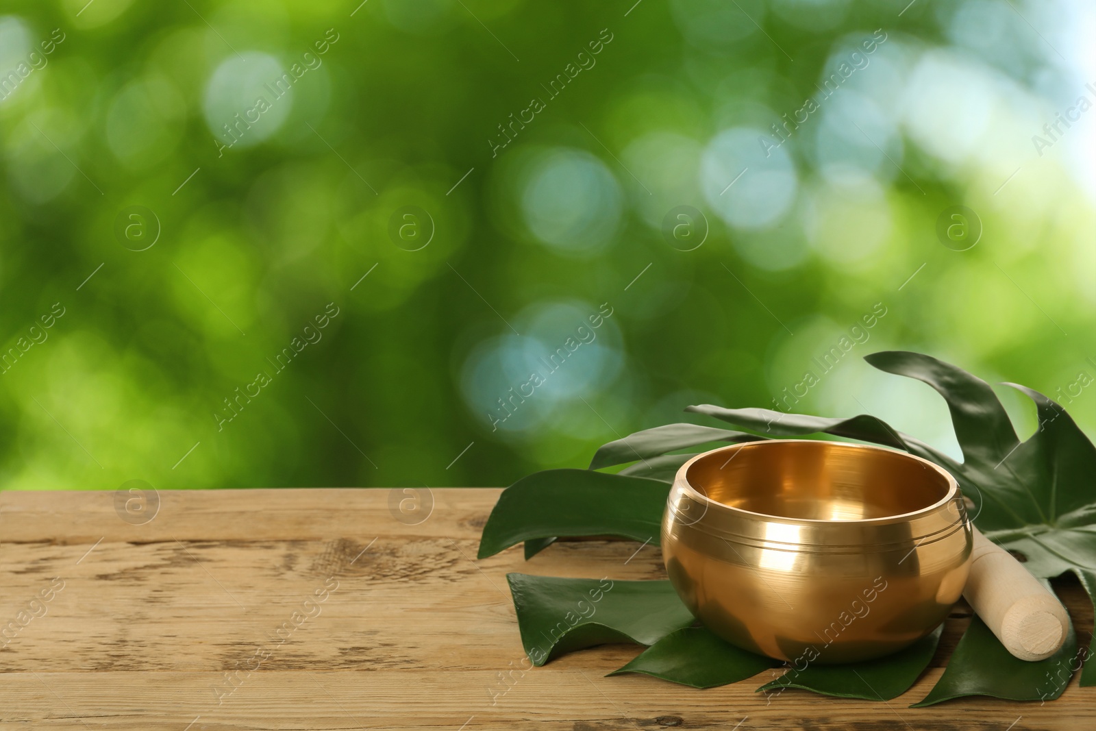 Image of Golden singing bowl, mallet and leaf on wooden table outdoors, space for text