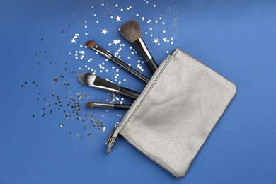 Different makeup brushes, case and shiny confetti on blue background, flat lay