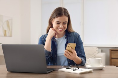 Photo of Happy woman with smartphone and laptop at wooden table in room