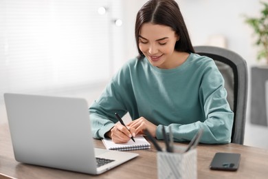 Photo of Young woman writing down notes during webinar at table in room