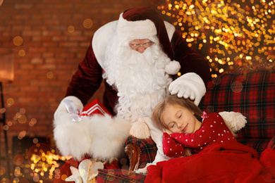 Photo of Santa Claus with Christmas gifts standing near sleeping little girl indoors